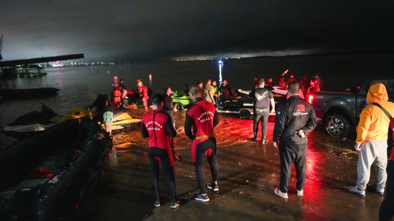 Foto noturna de várias pessoas envolvidas nos salvamentos próximas a um rio. Há embarcações de pequeno porte estacionadas. Algumas das pessoas usam trajes aquáticos.