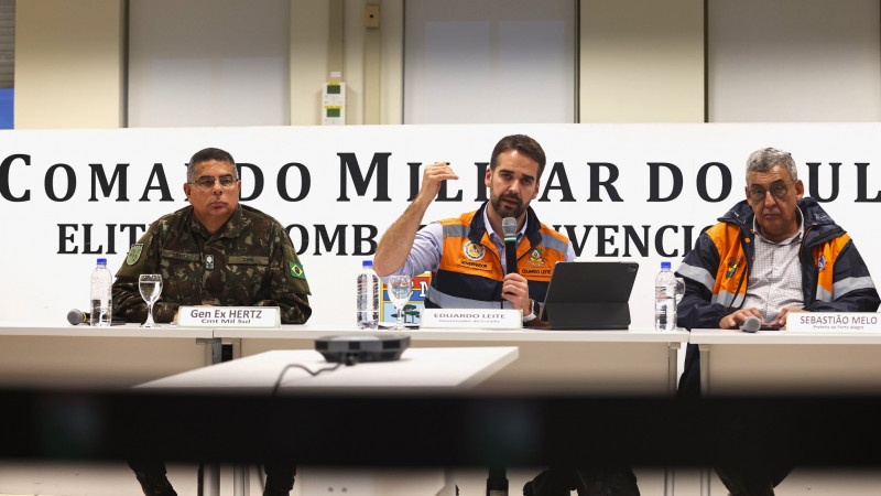 Leite, o prefeito de Porto Alegre e o coronel Hertz sentados atrás de uma mesa na coletiva sobre enchentes na sexta 3 de maio. Leite, com o microfone na mão, fala e gesticula. Atrás deles há um painel onde se lê Comando Militar do Sul.