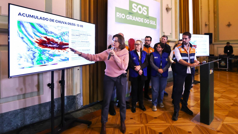 Meteorologista fala na coletiva sobre enchentes de 10 de maio de 2024. Ela aponta para uma tela com o mapa das chuvas enquanto Leite e outros secretários a observam.