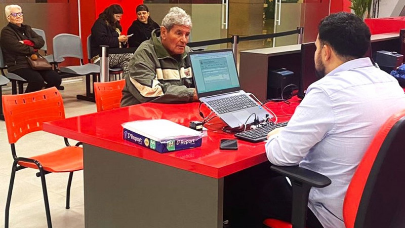 Tudo Fácil em ação para retirada de certidões após enchentes. Um home de costas trabalha em uma mesa com um computador e conversa com um cliente. Ao fundo, outros clientes aguardam sentados em cadeiras.