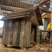 Foto enquadra casinha de madeira à esquerda, de lado. À direita, aparece um apenado da cintura pra baixo, de bermuda laranja e bota, martelando o telhado.