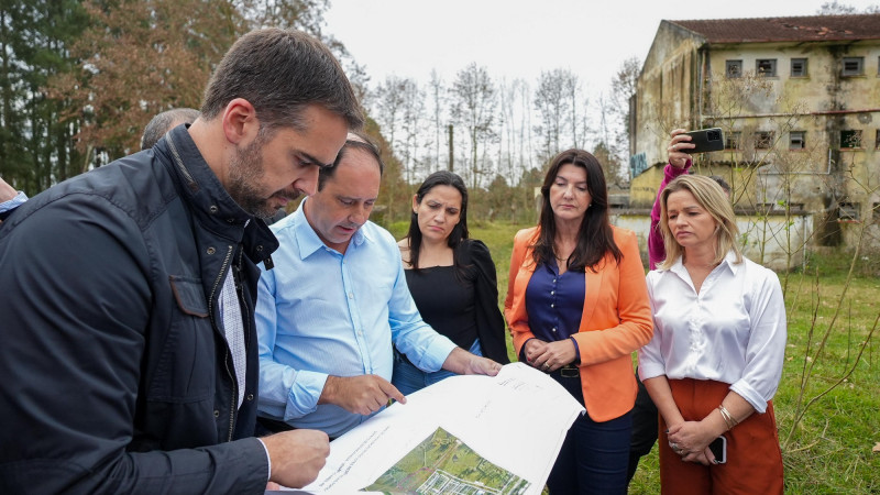 Leite anuncia construção de casas em Venâncio Aires. Na imagem ele olha em um papel detalhes do local onde as casas serão construídas. Outras observam o governador. Ao fundo, pode ser vista parte da edificação que será demolida para a construção das casas.