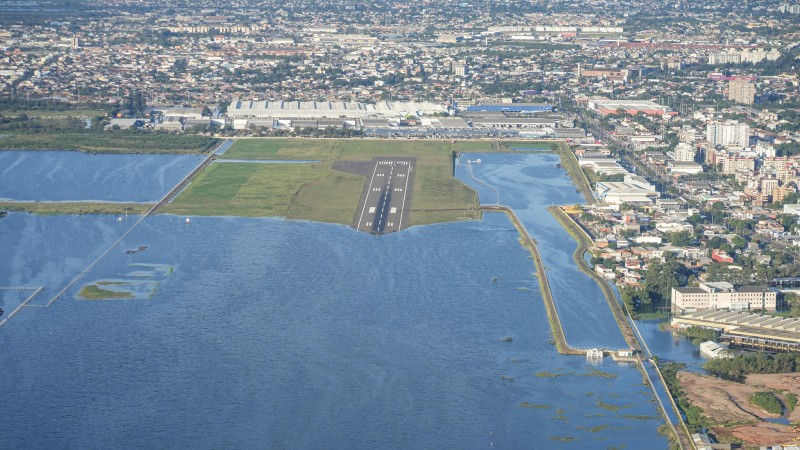 Imagem aérea do aeroporto alagado. Na parte inferior da foto aparece grande quantidade de água na pista. Ao fundo, a cidade de Porto Alegre.