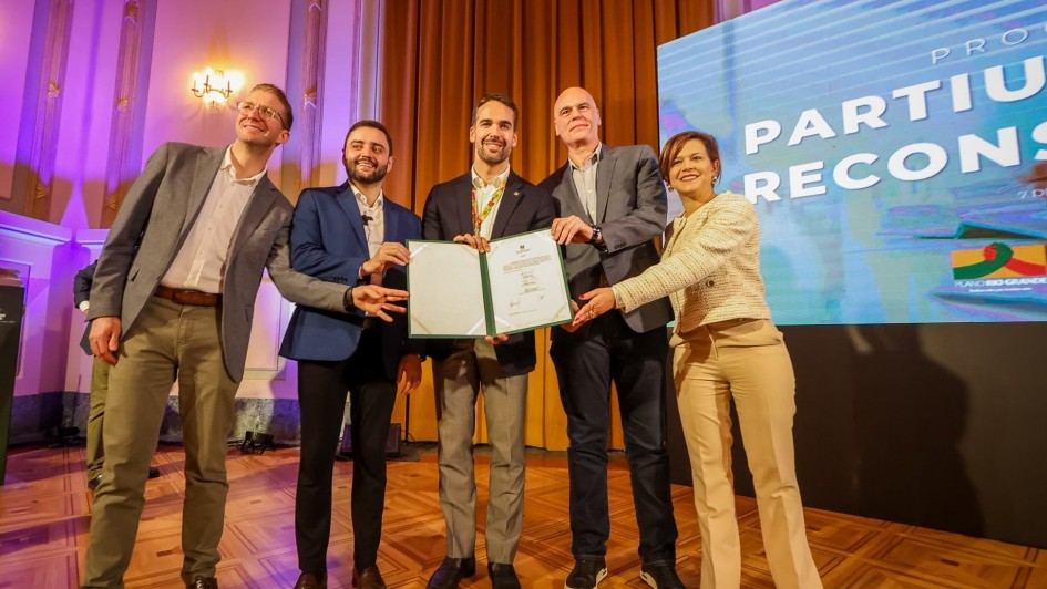 Leite e mais quatro pessoas segurando documento recém assinado no lançamento do Partiu Futuro Reconstrução. Todos estão em pé e trata-se de uma foto posada.