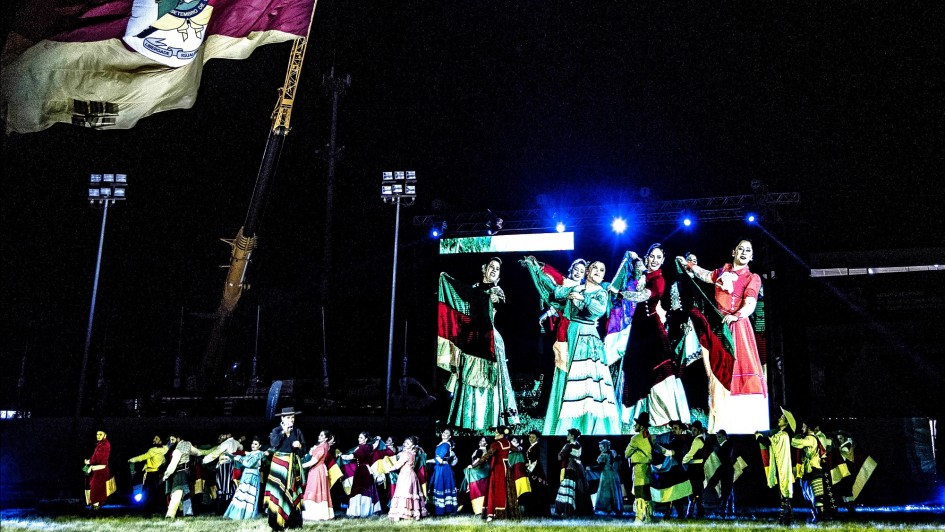 Imagem de uma apresenta&ccedil;&atilde;o anterior da &Oacute;pera Ga&uacute;cha na Expointer, com diversas pessoas dan&ccedil;ando e um enorme tel&atilde;o atr&aacute;s mostrando detalhes da dan&ccedil;a.