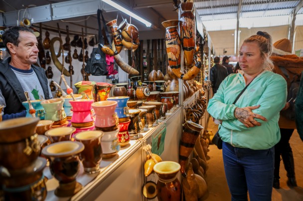 Estande com diversas cuias. Atrás do balcão está o artesão Kaiper. Na frente, uma mulher observa os produtos
