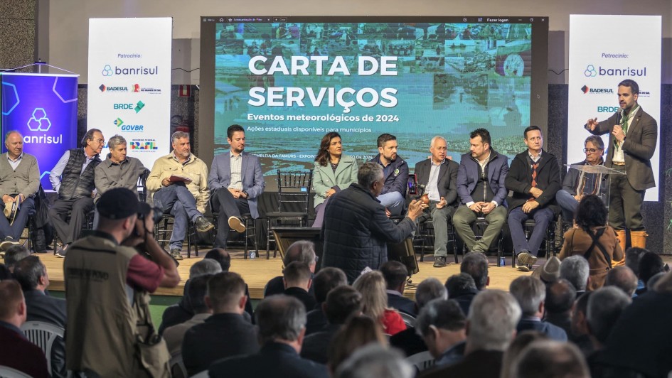 Leite ao microfone, em pé sobre um tablado falando para uma plateia que pode ser vista de costas. No tablado, há diversas pessoas sentadas acompanhando a fala do governador.