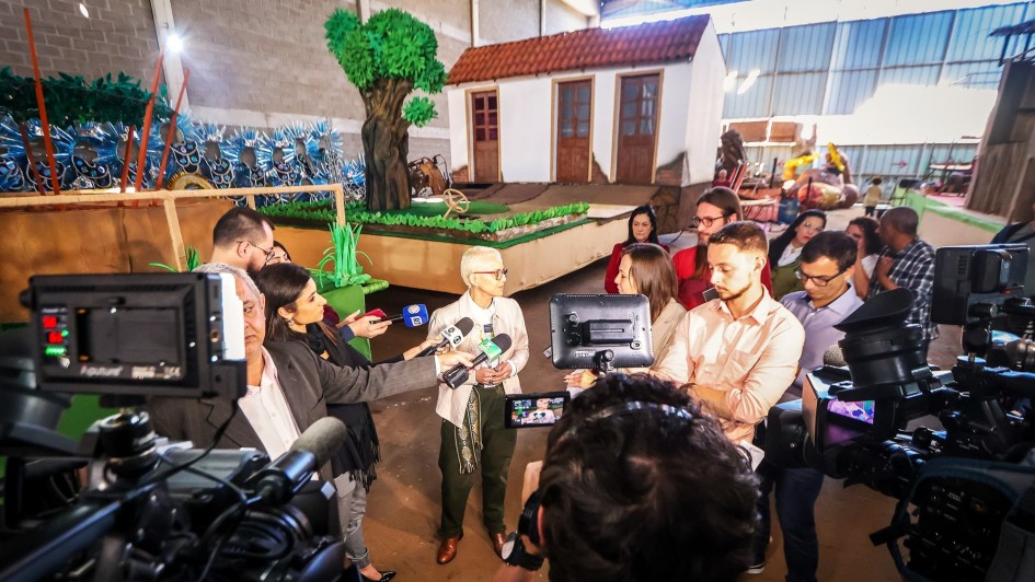 Beztriz Araujo rodeado de jornalistas, câmeras e microfones dando entrevista. Ao fundo, há uma alegoria do desfile já preparada para o desfile.