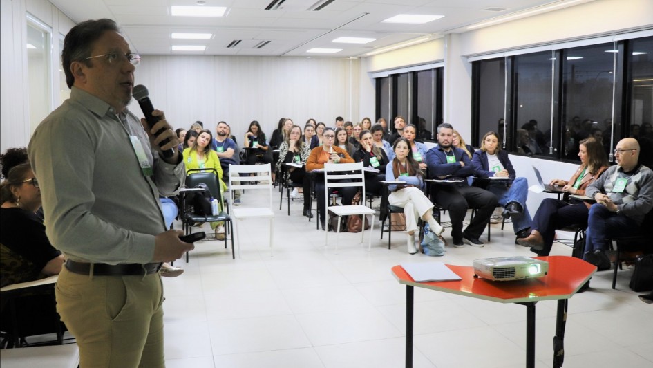 À esquerda um professor em pé explicando algo e nos restante da imagem os alunos do curso, sentados em carteiras, assistindo à aula.