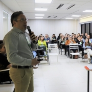 À esquerda um professor em pé explicando algo e nos restante da imagem os alunos do curso, sentados em carteiras, assistindo à aula.