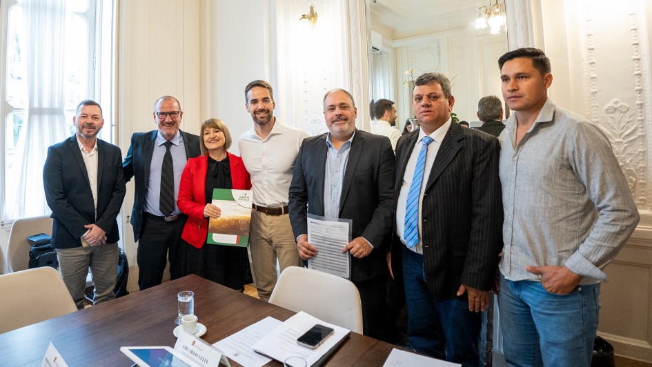 A imagem mostra sete pessoas em pé, incluindo Leite e Rosemar, posando para uma foto em um ambiente formal. O ambiente é elegante, com paredes claras e detalhes decorativos clássicos. Há uma mesa com papéis e um tablet à frente do grupo.