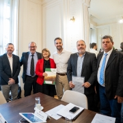 A imagem mostra sete pessoas em pé, incluindo Leite e Rosemar, posando para uma foto em um ambiente formal. O ambiente é elegante, com paredes claras e detalhes decorativos clássicos. Há uma mesa com papéis e um tablet à frente do grupo.