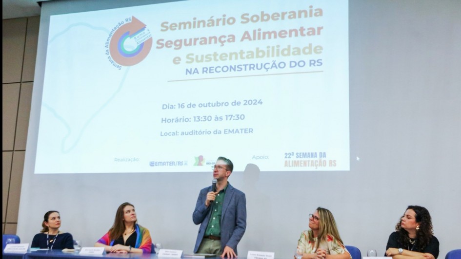 Um homem em pé falando ao microfone sobre um palco e atrás da mesa do evento sendo observado por quatro mulheres que também compõem a mesa da Semana da Alimentação RS 2024.