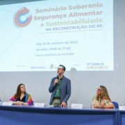 Um homem em pé falando ao microfone sobre um palco e atrás da mesa do evento sendo observado por quatro mulheres que também compõem a mesa da Semana da Alimentação RS 2024.