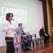 Raquel em pé, atrás de um púlpito e falando ao microfone sobre o placo no Seminário de Carreira Docente 2024. Ela é observada por quatro homens que compõem a mesa de abertura do evento.