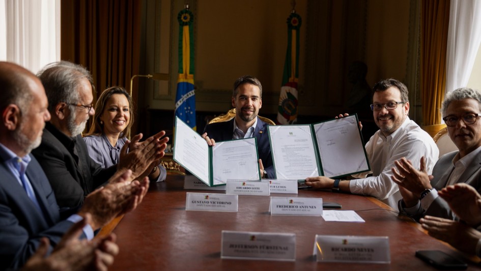 Foto posada em que Leite e Lemos seguram o documento recém assinado. Eles estão sentados à mesa de reunião rodeados de autoridades.