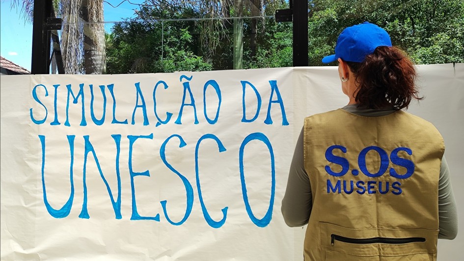 Imagem de uma pessoal de costas com um colete escrito SOS Museus fixando uma faixa com os dizeres: Simulação da Unesco.