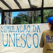 Imagem de uma pessoal de costas com um colete escrito SOS Museus fixando uma faixa com os dizeres: Simulação da Unesco.