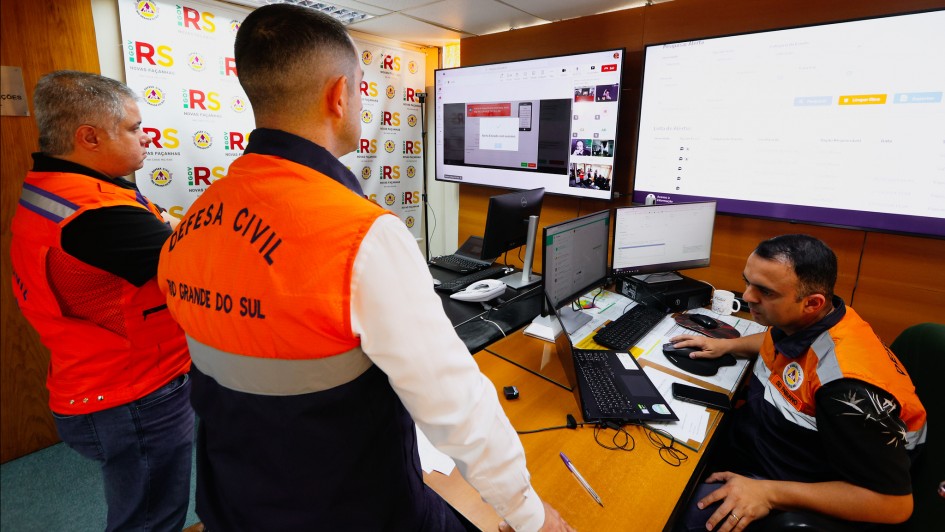 Três pessoas em uma sala de controle. Duas delas observam uma terceira, que está sentada diante de um computador realizando o envio da mensagem.