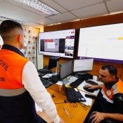 Três pessoas em uma sala de controle. Duas delas observam uma terceira, que está sentada diante de um computador realizando o envio da mensagem.