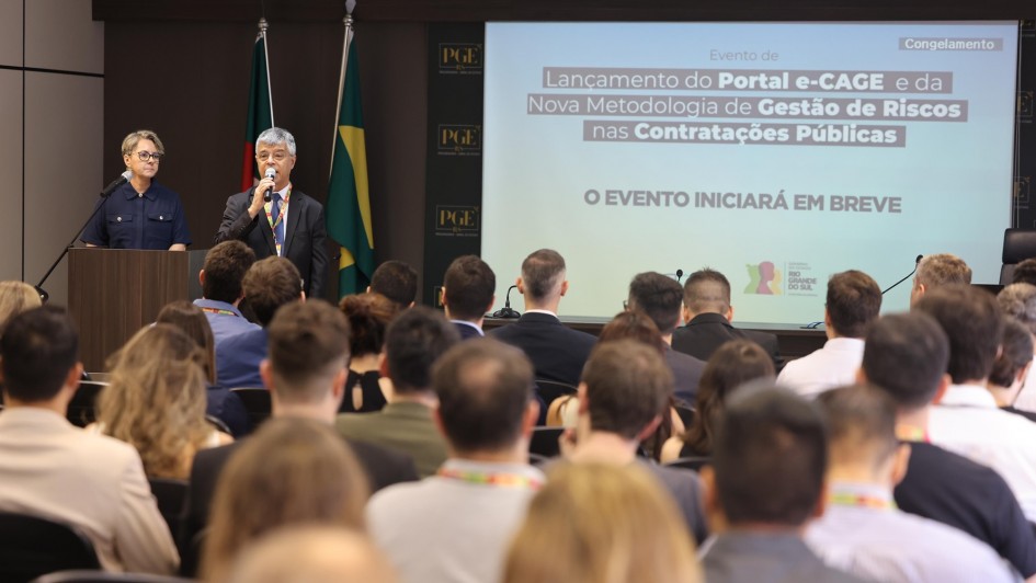 Antônio Kehrwald falando ao microfone para a plateia que pode ser vista de costas na imagem.