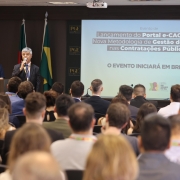 Antônio Kehrwald falando ao microfone para a plateia que pode ser vista de costas na imagem.