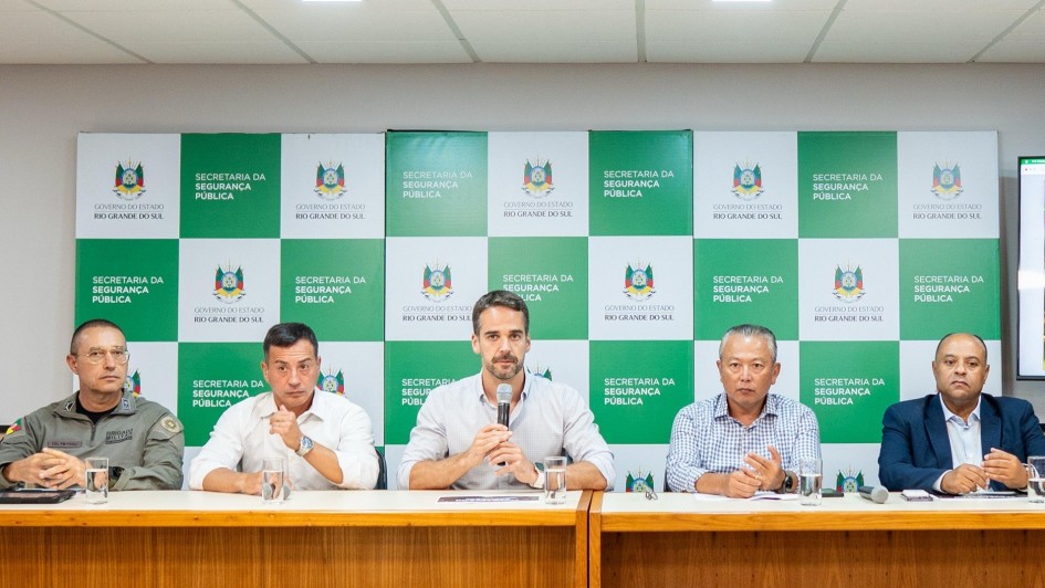 Leite e mais quatro homens sentados atrás de uma bancada para a coletiva. Leite segura o microfone e fala com os jornalistas.