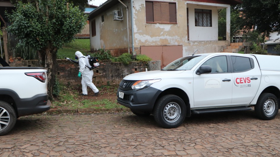 Numa rua residencial de pedra, um agente de saúde com roupa de proteção branca cobrindo todo o corpo caminha com o equipamento de inseticida nas costas. Há um carro branco do Cevs estacionado.