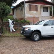 Numa rua residencial de pedra, um agente de saúde com roupa de proteção branca cobrindo todo o corpo caminha com o equipamento de inseticida nas costas. Há um carro branco do Cevs estacionado.