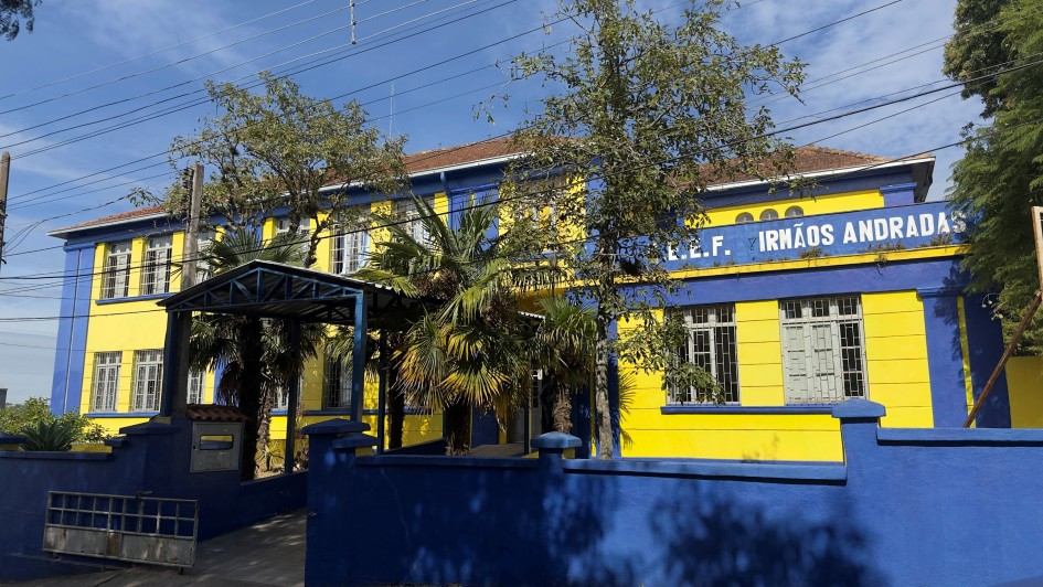 Foto de uma escola nas cores azul e amarelo. Há algumas árvores na entrada da escola, na parte de dentro. Aparece o céu azul.