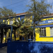 Foto de uma escola nas cores azul e amarelo. Há algumas árvores na entrada da escola, na parte de dentro. Aparece o céu azul.