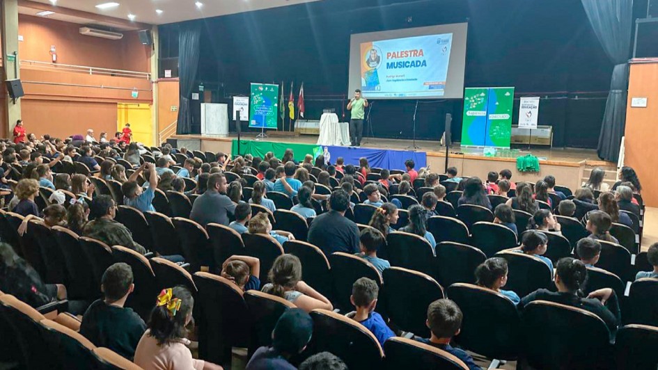 Em um auditório há dezenas de crianças sentadas assistindo uma apresentação. No palco há um palestrante e três banners. O fundo tem cortinas escuras e do lado esquerdo há uma parede marrom.