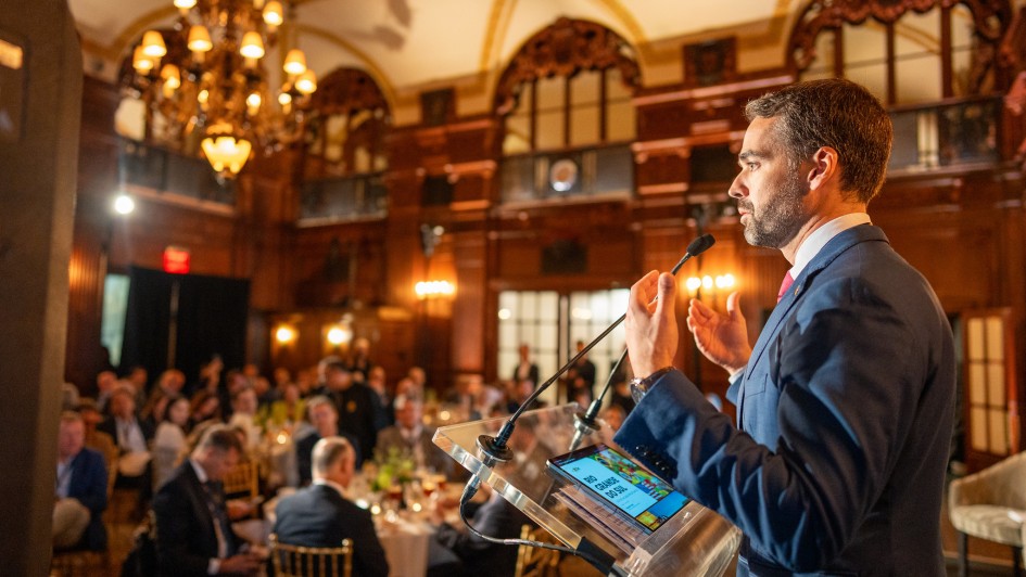 Leite ao microfone gesticulando durante fala no RS Day 2025 em Nova York. A parte da plateia pode ser vista sentada em mesas de almoço.