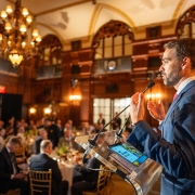 Leite ao microfone gesticulando durante fala no RS Day 2025 em Nova York. A parte da plateia pode ser vista sentada em mesas de almoço.
