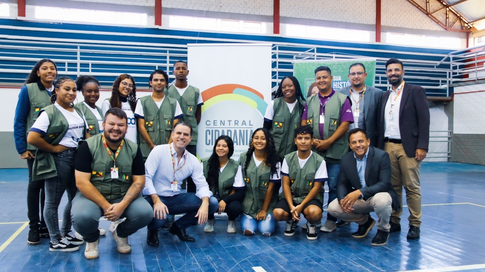 Há um grupo de pessoas reunidos para tirar foto em um ginásio de piso azul e com um cartaz ao fundo.