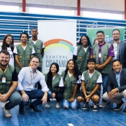Há um grupo de pessoas reunidos para tirar foto em um ginásio de piso azul e com um cartaz ao fundo. 