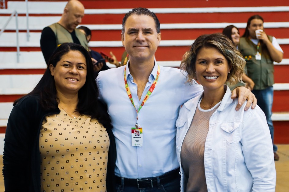 Há três pessoas de pé sorrindo e posando para foto. Duas mulheres e um homem. Eles estão abraçados. O homem usa crachá. Ao fundo há três pessoas de colete de pé e uma escadaria nas cores branca e vermelha.