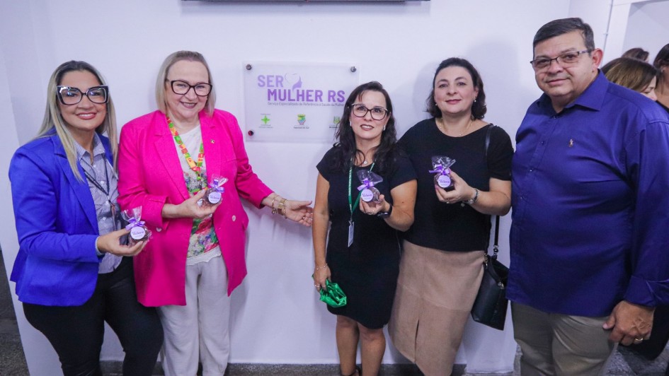 Há cinco pessoas posando para uma foto. Quatro usam óculos. Um é homem. Ao fundo tem um painel escrito SER Mulher RS. As quatro mulheres seguram nas mãos uma espécie de medalhinha.