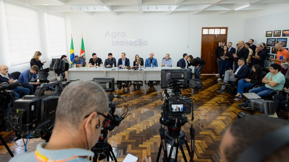 Imagem geral de coletiva sobre gripe aviária em maio de 2025. Na mesa diversas pessoas ligadas ao governo e de costas e nas laterais jornalistas assitiindo.