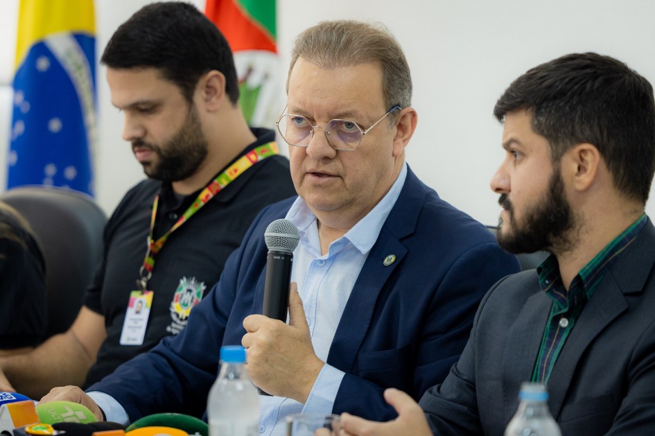 Secretário da Agricultura em close falando ao microfone do lado de dois homens na coletiva sobre gripe aviária em maio de 2025