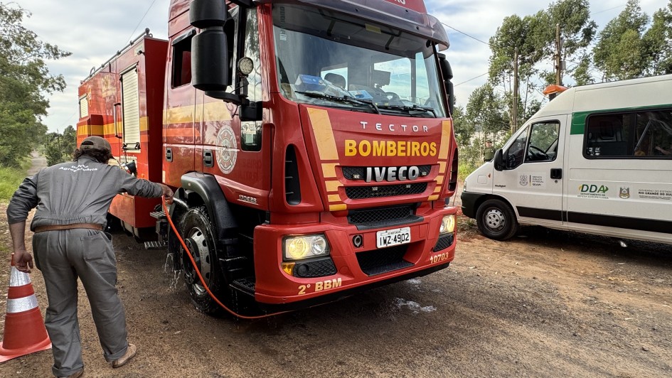Caminhão dos bombeiros militar parado em barreira sanitária e homem lava com água as rodas do veículo dos bombeiros em estrada rural. Ao fundo, à direita, aparece uma van branca do Departamento de Vigilância e Defesa Sanitária Animal da Secretaria da Agricultura, Pecuária, Produção Sustentável e Irrigação (DDA/Seapi)
