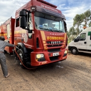 Caminhão dos bombeiros militar parado em barreira sanitária e homem lava com água as rodas do veículo dos bombeiros em estrada rural. Ao fundo, à direita, aparece uma van branca do Departamento de Vigilância e Defesa Sanitária Animal da Secretaria da Agricultura, Pecuária, Produção Sustentável e Irrigação (DDA/Seapi)