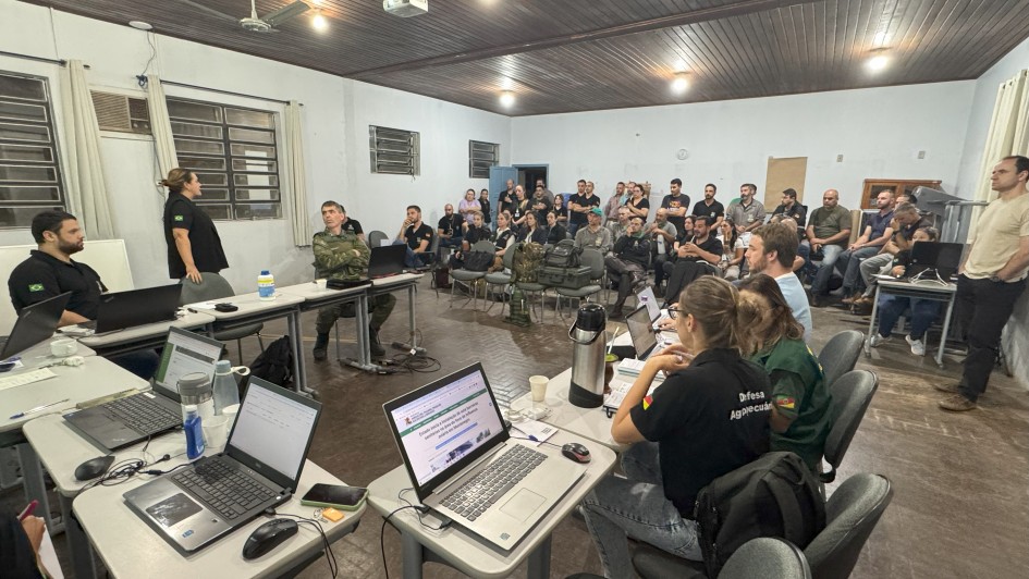 A foto mostra um grupo de pessoas reunidas em uma sala. H&aacute; mesas com laptops e pessoas sentadas, algumas olhando para a frente e outras trabalhando em seus computadores.