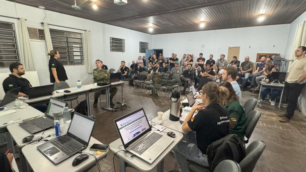 A foto mostra um grupo de pessoas reunidas em uma sala. Há mesas com laptops e pessoas sentadas, algumas olhando para a frente e outras trabalhando em seus computadores.