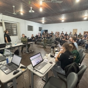 A foto mostra um grupo de pessoas reunidas em uma sala. Há mesas com laptops e pessoas sentadas, algumas olhando para a frente e outras trabalhando em seus computadores.