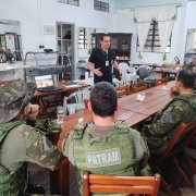 A foto mostra um homem de camisa preta gesticula em frente a um grupo de pessoas fardadas sentadas em mesas de madeira, algumas olhando para um laptop aberto.