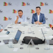 Há 8 pessoas sentadas. Em uma mesa retangular, há notebooks, celulares e copo de água. Três pessoas estão de terno. Ao fundo há um painel do Estado do Rio Grande do Sul.