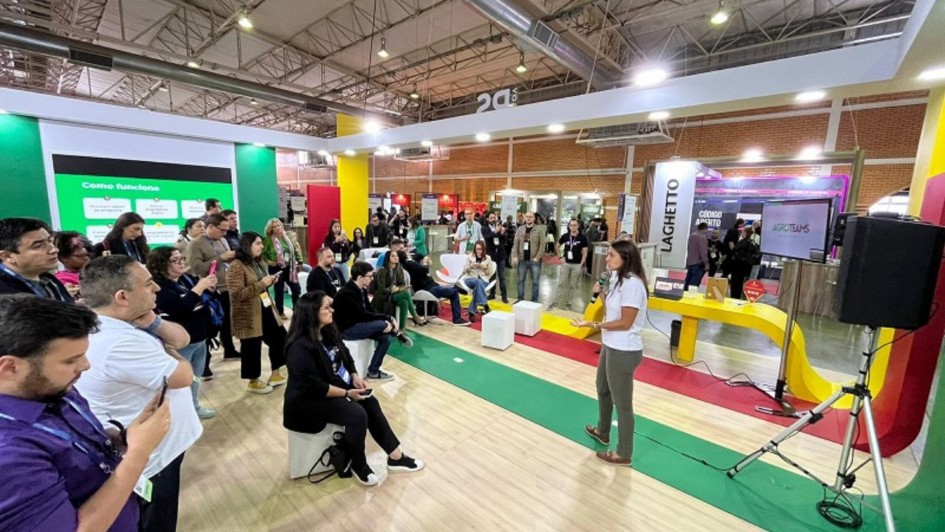 Há dezenas de pessoas, algumas sentadas e a maioria de pé, escutando uma palestrante. Ela segura um microfone. O chão tem cor clara e partes em verde, vermelho e amarelo. Ao fundo há um painel de apresentação.