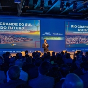 Em um palco de apresentação, há um homem de pé segurando um microfone. Dezenas de pessoas sentadas assistem ele. O palco tem a cor azul e há dois painéis grandes escrito Rio Grande do Sul do Futuro.