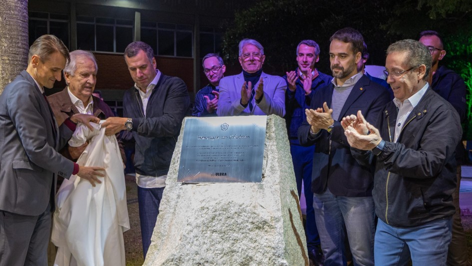 Quatro homens revelam uma placa comemorativa em uma pedra rústica, enquanto outras autoridades e convidados aplaudem ao fundo, em evento noturno.